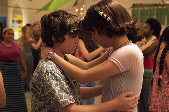 A boy and girl in casual clothing dance closely together in a crowded, warm-toned room.
