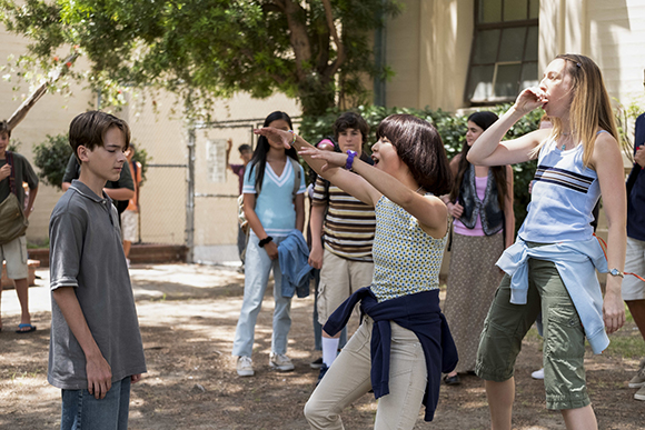 Several teenagers stand in a courtyard; one girl yells while another reaches out toward a boy.