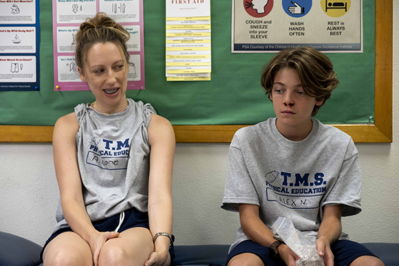 A woman in a grey tank top and a teenage boy in a grey t-shirt sit on a blue bench in front of a green bulletin board.