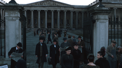 People in early 20th-century attire walk through large gates toward a building with a grand colonnade.