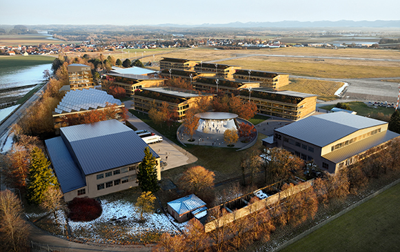 Aerial view of a large modern building complex with flat roofs, surrounded by autumn trees and open fields.