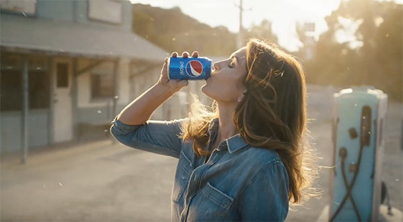 A woman in a denim shirt drinks from a blue Pepsi can outdoors during sunset.