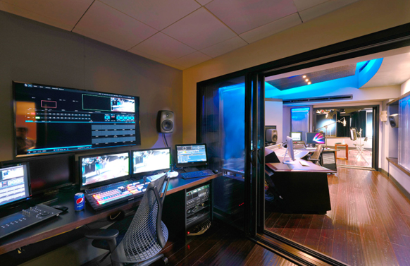 A video editing workstation with several monitors, a mixing console, and a view into a blue-lit studio.
