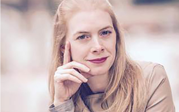 A woman with long blonde hair looks at the camera while resting her chin on her hand.