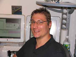 A smiling man with glasses sitting in front of two computer monitors in an office setting.