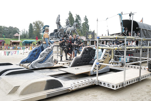 Film crew using cameras and lighting equipment to film a person in a chariot on a sandy outdoor set.