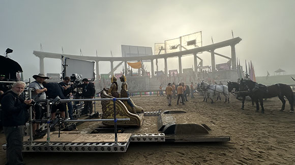 Film crew with cameras and equipment on a sandy set near a large structure and horse-drawn carriages in fog.