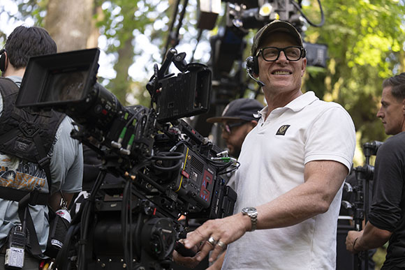 A man in a white polo and glasses smiles while operating professional film camera equipment outdoors.