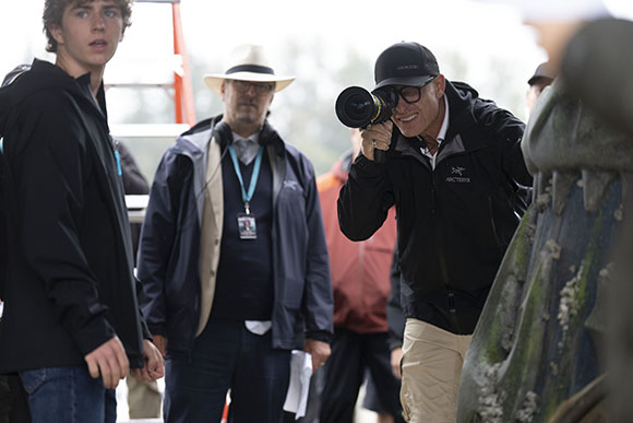 A man in a black jacket and cap looks through a camera lens while others stand nearby.