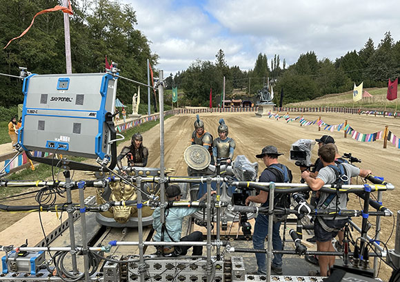Film crew using a camera rig and lighting equipment on a dirt track set with actors in costume.