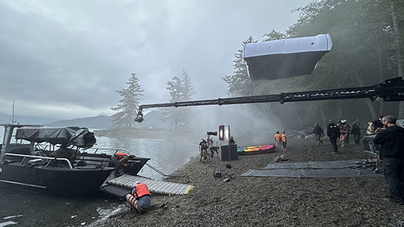 A camera crane extends over a lake near a boat and a film crew on a pebbly shore under a foggy sky.