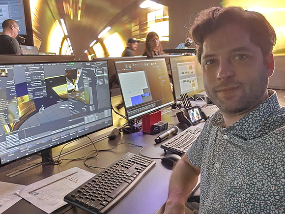 A man in a patterned shirt takes a selfie in a dark control room filled with computer monitors.