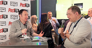 Two men sit at a table for an interview in front of a backdrop with logos.