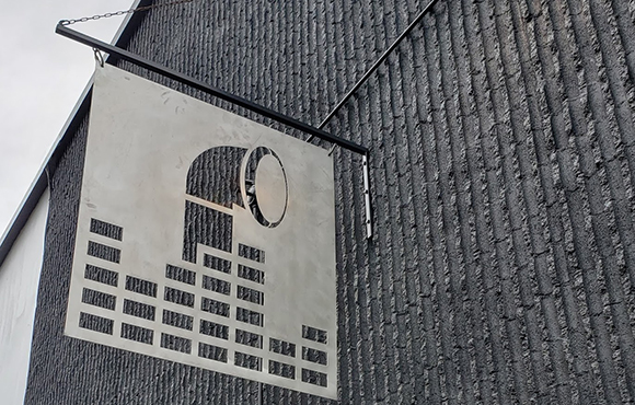 A rectangular metal sign with a cutout design of a building and a horn hangs from a dark, textured wall.