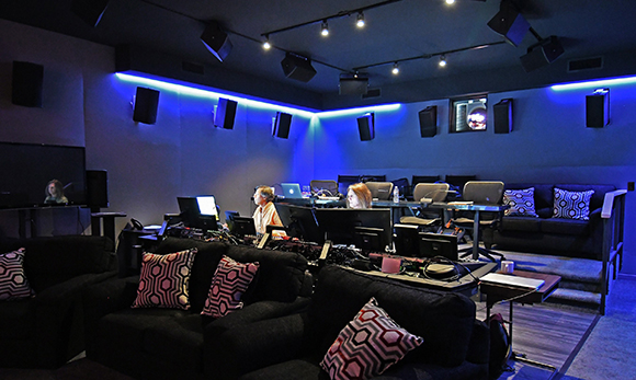 A dark sound mixing studio with blue neon lighting, multiple monitors, speakers, and black sofas.