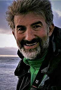 A smiling man with salt-and-pepper hair and a beard wearing a green shirt and black jacket.
