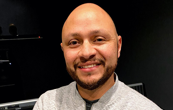A smiling bald man with a dark beard wearing a grey zip-up sweatshirt against a dark background.