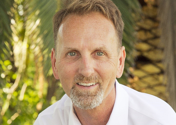 A smiling middle-aged man with light eyes and a graying goatee wearing a white shirt.