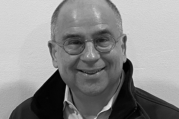 Black and white close-up portrait of a smiling man wearing round glasses and a dark jacket.