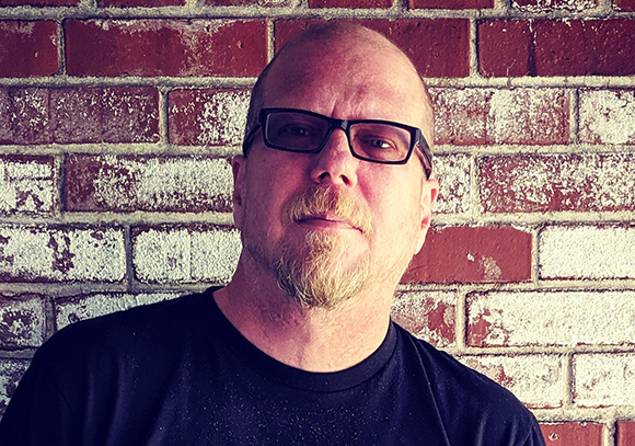 A man with glasses and a goatee wearing a black shirt against a red and white brick wall.