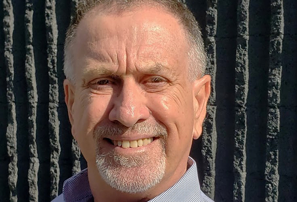 A smiling middle-aged man with short hair and a grey goatee in front of a dark, vertically ribbed wall.