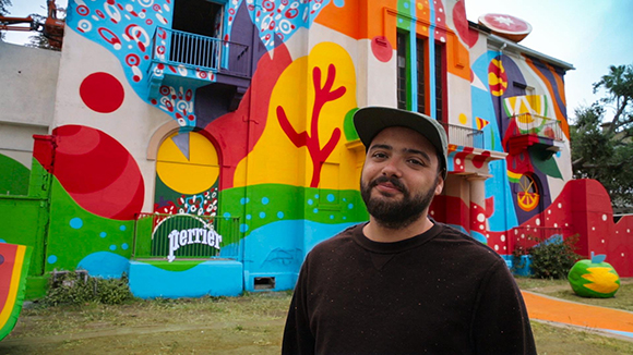 A man with a beard and cap stands before a building covered in vibrant, multi-colored abstract murals.