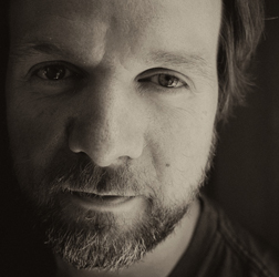 A close-up, sepia-toned portrait of a man with a beard looking directly at the camera.