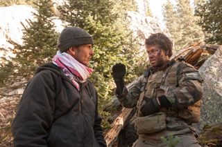 A man in a dark jacket and pink scarf talks to a man in camouflage gear in a forest.