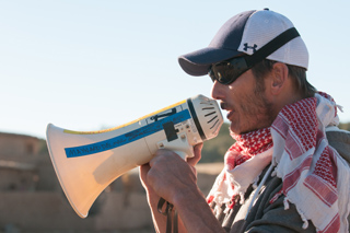 A man in a baseball cap and sunglasses holds a white megaphone to his mouth.