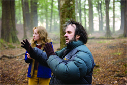 A man in a dark puffer jacket gestures with gloved hands while a woman stands behind him in a wooded area.