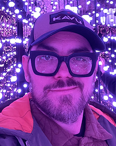 A man with a beard, glasses, and a KAVU cap smiling in front of bright purple lights.