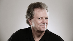 A middle-aged man with wavy brown hair wearing a black shirt, smiling against a neutral background.