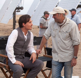 A man in a grey vest sits in a director's chair talking to a man in a tan shirt and baseball cap.