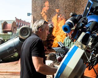 A man in a black shirt looks toward a large fire behind a professional film camera rig.