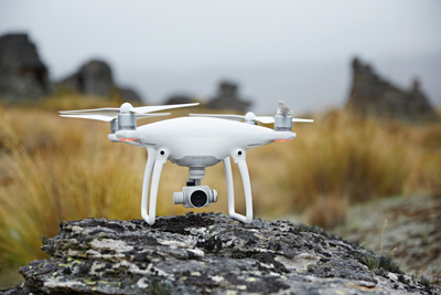 A white quadcopter drone with a camera gimbal sits on a large, lichen-covered rock in a grassy field.