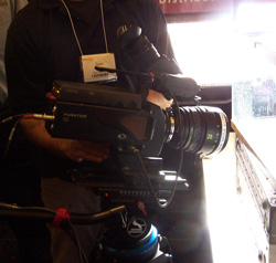 A person in a black shirt holds a professional video camera with a large lens and a tag on their shirt.