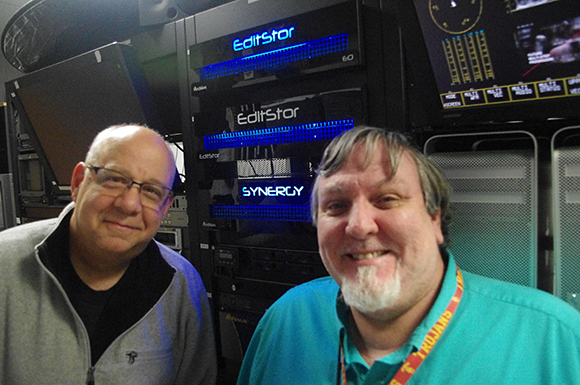 Two men smiling in front of black EditStar and Synergy server equipment.