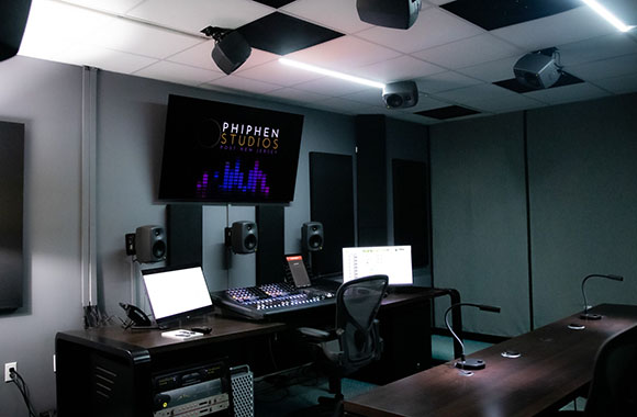 A dark recording studio featuring a large screen displaying Phiphen Studios logo, mixing consoles, and studio monitors.