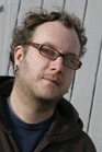 A man with curly hair, glasses, and a goatee wearing a dark hoodie.