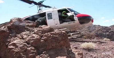 A white helicopter with red accents hovers closely above a steep, rocky mountain ridge.