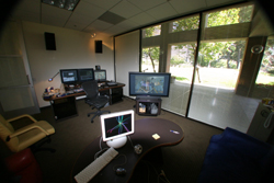 An office with several computer monitors, desks, and large windows overlooking trees.