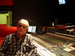 A man in a plaid shirt sits in a dark recording studio behind a large audio mixing console.