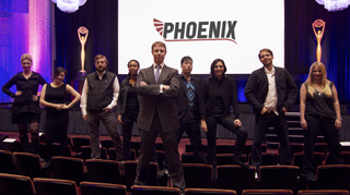 A group of seven people posing on a stage in front of a large white screen with the word PHOENIX.