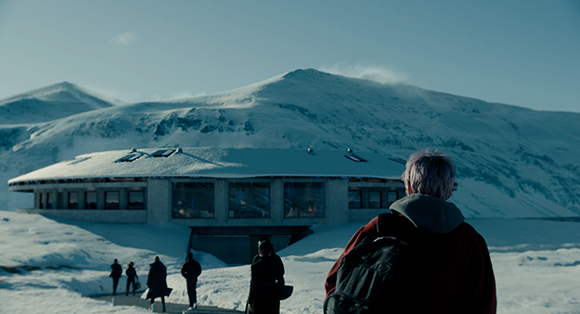 A person with a backpack looks at a large, low-profile circular building in a snowy mountain range.