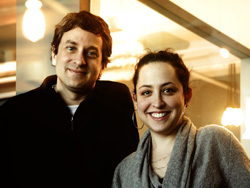 A man in a black jacket and a woman in a grey sweater smiling side by side.