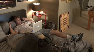 A young man with a prosthetic leg lies in bed using a laptop while talking on a phone.