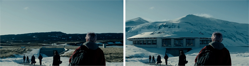A person with a backpack looks toward a snowy mountain landscape and a large, low-profile building.