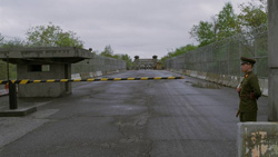 A soldier in uniform stands near a yellow and black striped barrier at a checkpoint.