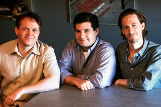 Three men sitting side by side at a dark table against a blue wall.