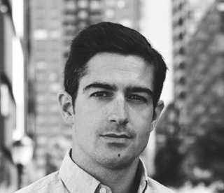 Black and white headshot of a man with dark hair wearing a button-down shirt against a blurred city background.